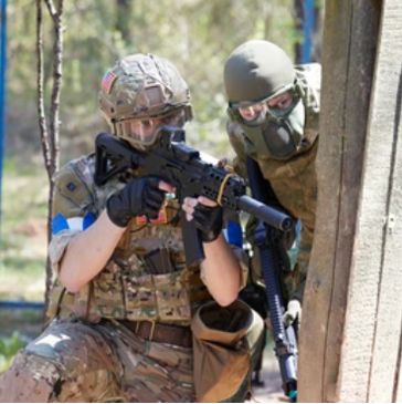 Two individuals in tactical gear aiming rifles behind cover outdoors.