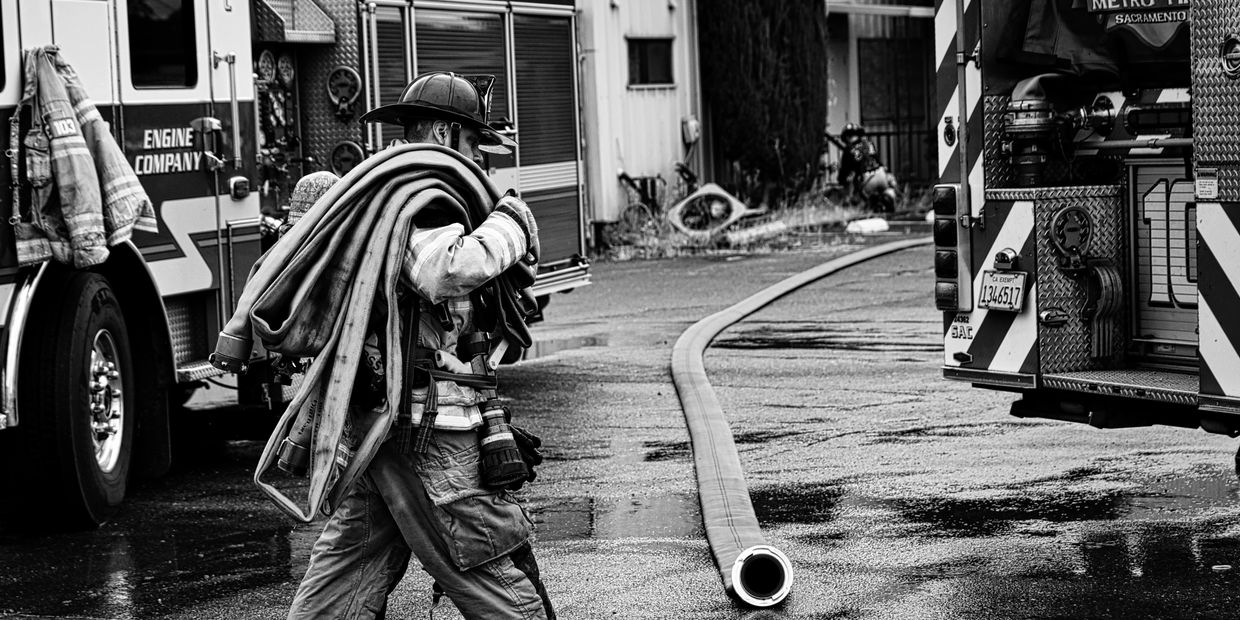 Firefighter carrying a coiled hose near fire trucks on a wet street.