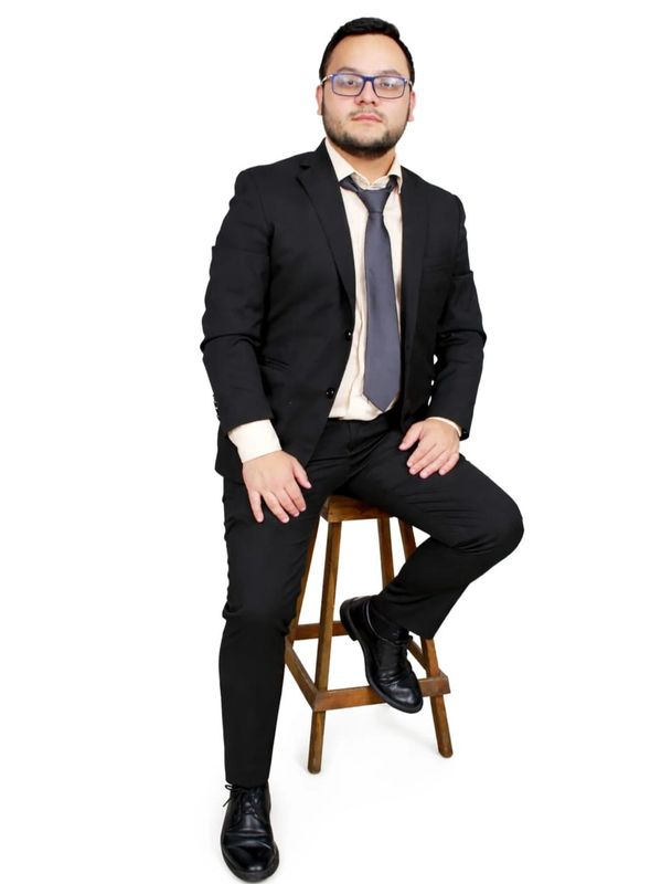 Man in black suit sitting on a wooden stool against white background.