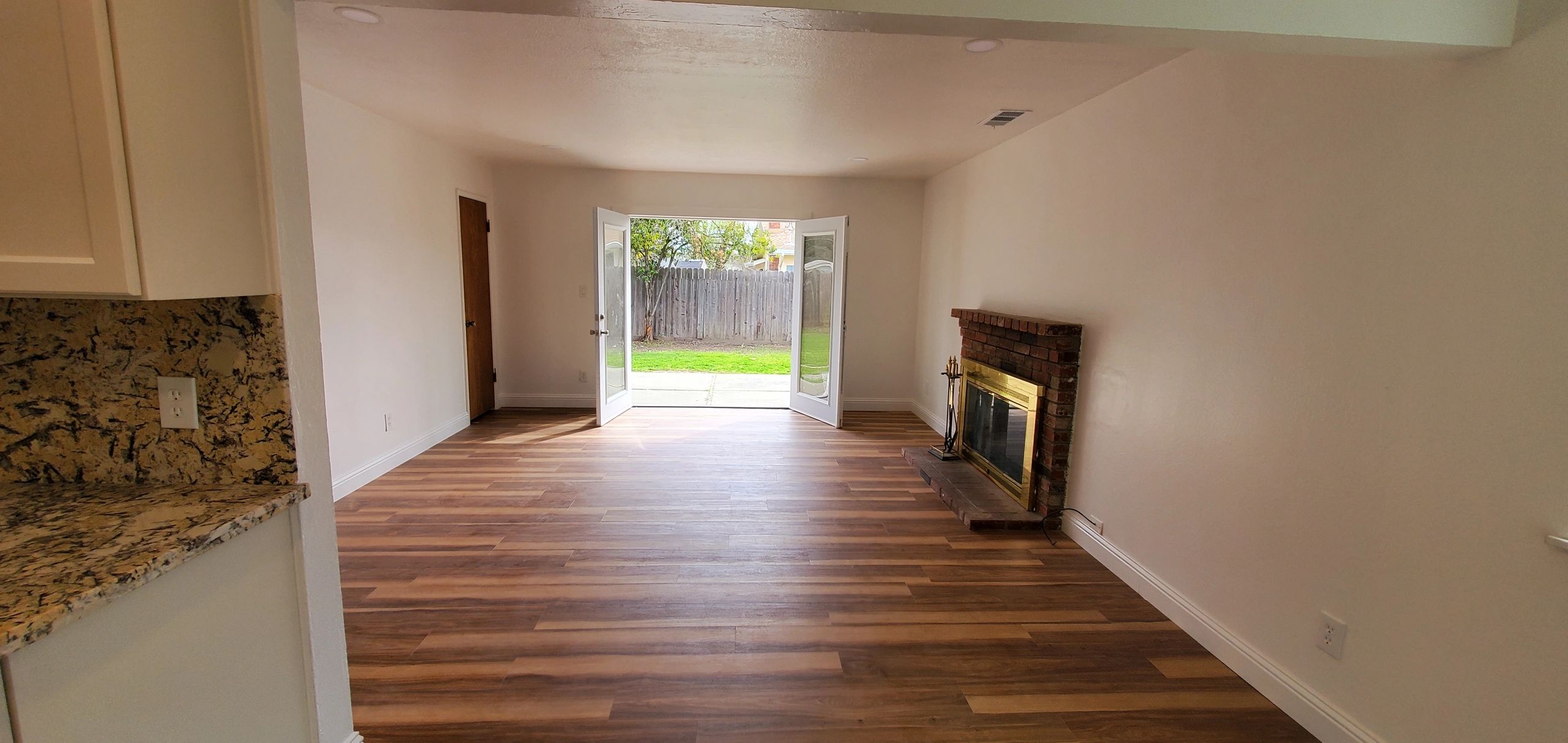 Empty living room with wooden floors, brick fireplace, and open doors to a backyard.