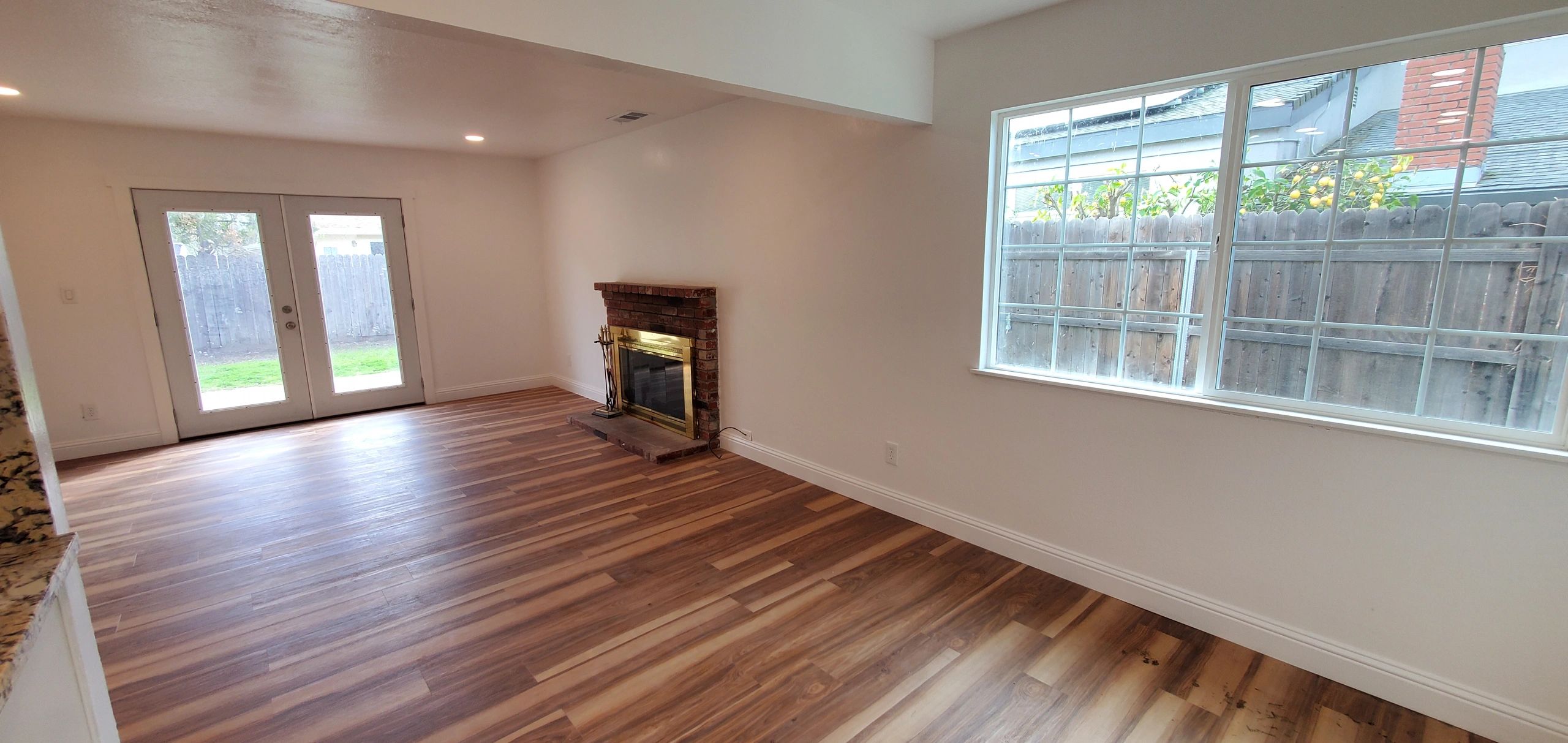 Empty living room with wooden floor, large window, and brick fireplace.