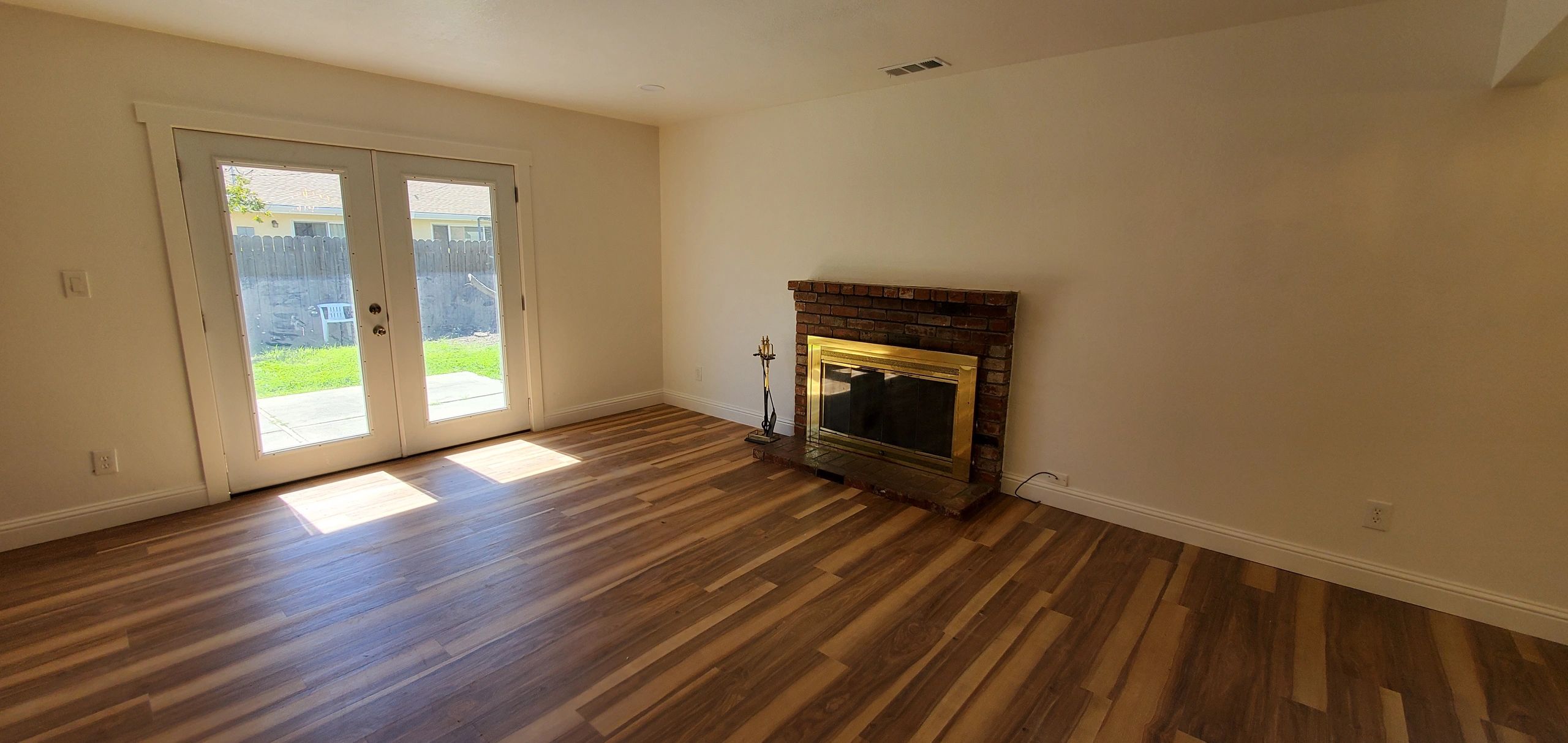 Empty room with wooden floor, brick fireplace, and glass doors to backyard.