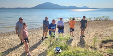 A group of six people standing on a lakeshore with mountains in the background.
