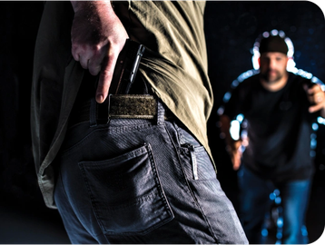 A person pulling a gun from their pants pocket with a blurred figure approaching.