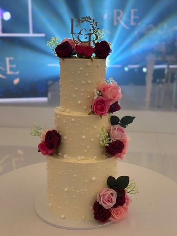 Elegant three-tier wedding cake adorned with red and pink roses and initials topper.