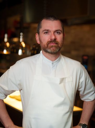 Confident male chef in a white apron standing in a warmly lit kitchen.