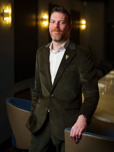 Man in a dark green suit standing by a dining table in a dimly lit room.