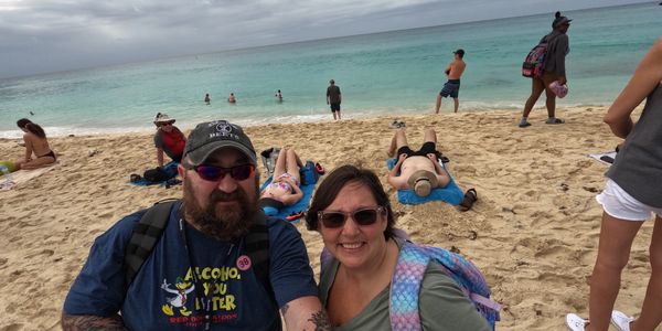 Wayne and Rena smiling on the beach in St. Martin with turquoise water and a mix of sunbathers.