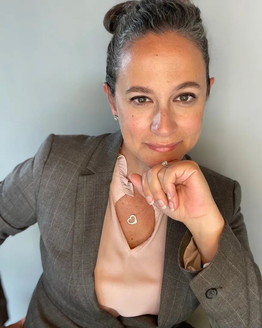 Confident woman in a suit with a heart necklace and thoughtful expression.