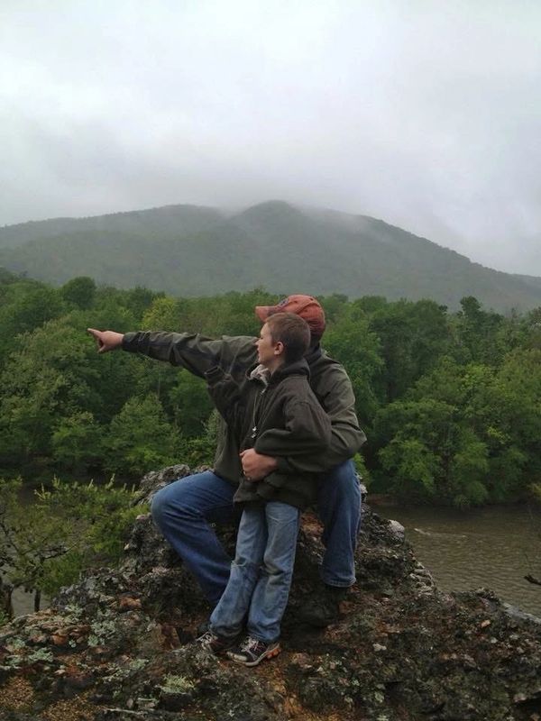 A man and boy on a rock overlooking a forested river valley.