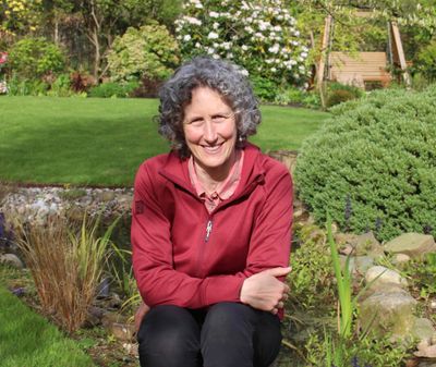 A smiling woman with curly gray hair sitting in a garden on a sunny day.
