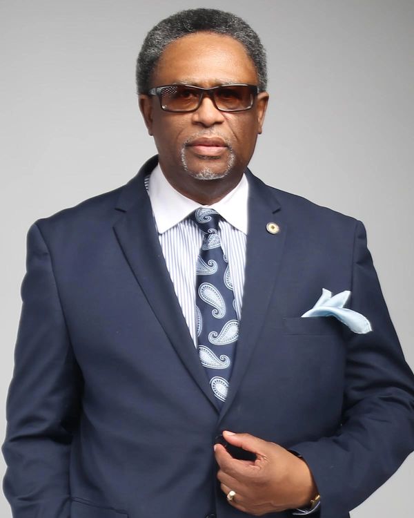 Man in a navy suit with patterned tie and sunglasses.