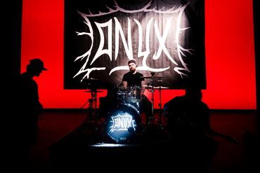 A drummer performs on stage with a bright red background and a band's logo.