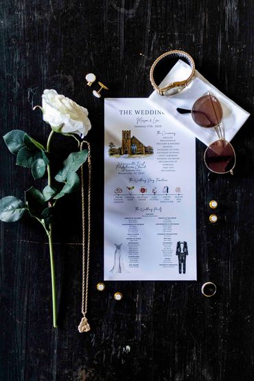 Wedding invitation with accessories and a white rose on a dark surface.