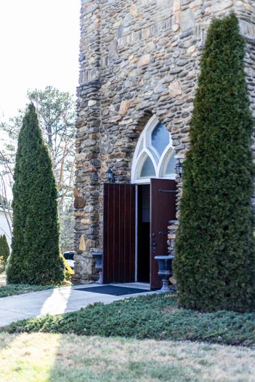 Stone building with arched window and open wooden door between two tall conical shrubs.