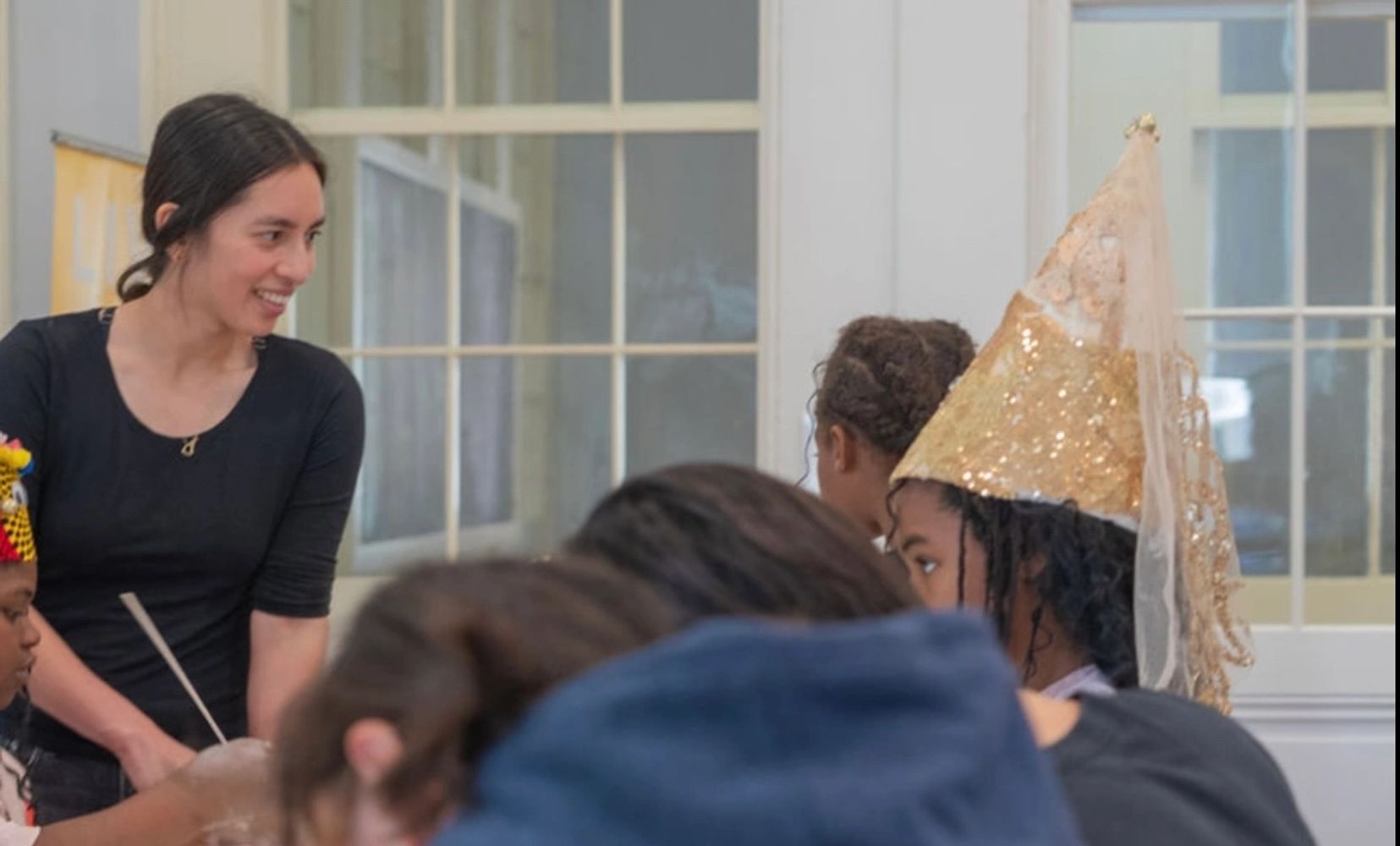 A woman in black engaging with children, one wearing a sparkling golden cone hat.
