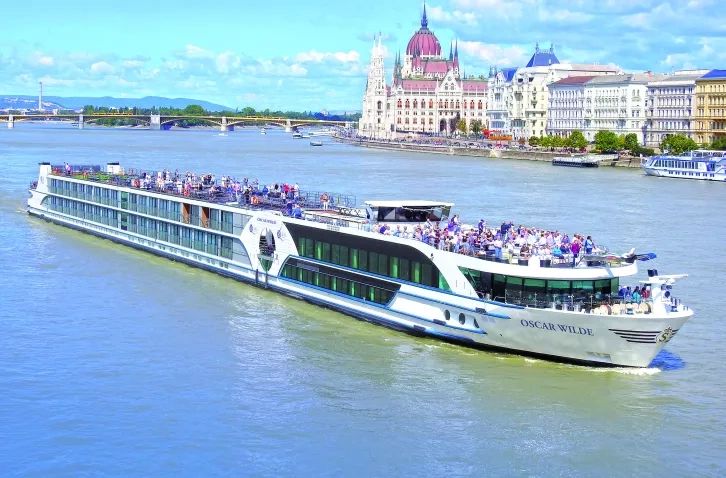A large river cruise ship named Oscar Wilde sailing on a river near a historic city.