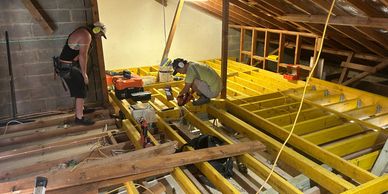 Two workers installing yellow wooden floor joists in an attic under construction.