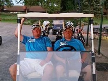 Two men in blue shirts sitting in a golf cart with golf clubs behind them.