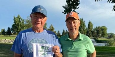 Two men outdoors holding a seniors golf championship sign.