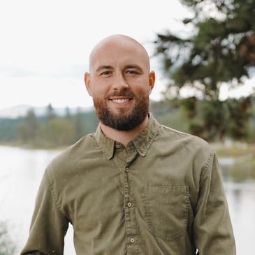 Smiling bald man with a beard wearing an olive green shirt outdoors by a lake.