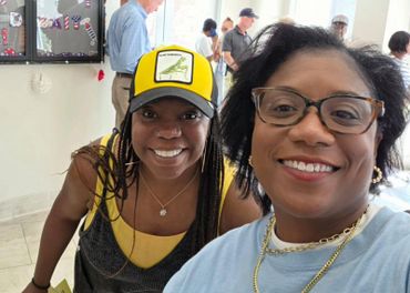 Two smiling women, one wearing glasses and the other a yellow cap, taking a selfie indoors.