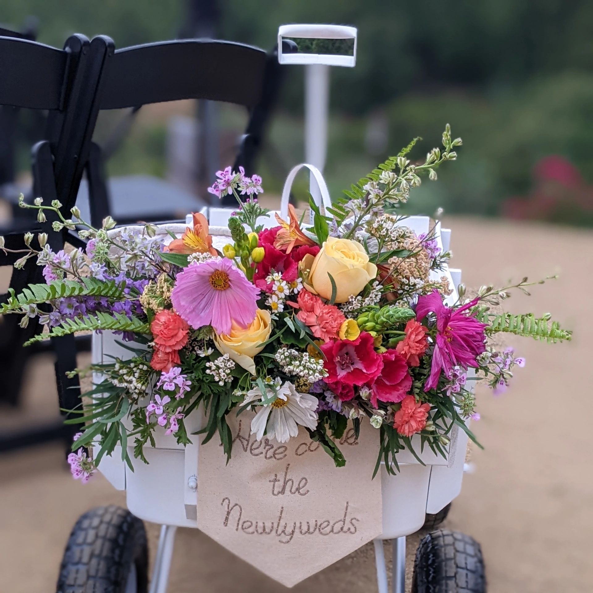 Colorful wedding flower arrangement on a cart with 'Here come the Newlyweds' sign.