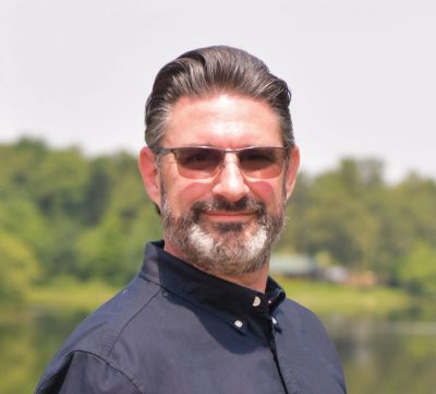 Man with glasses smiling outdoors near water.