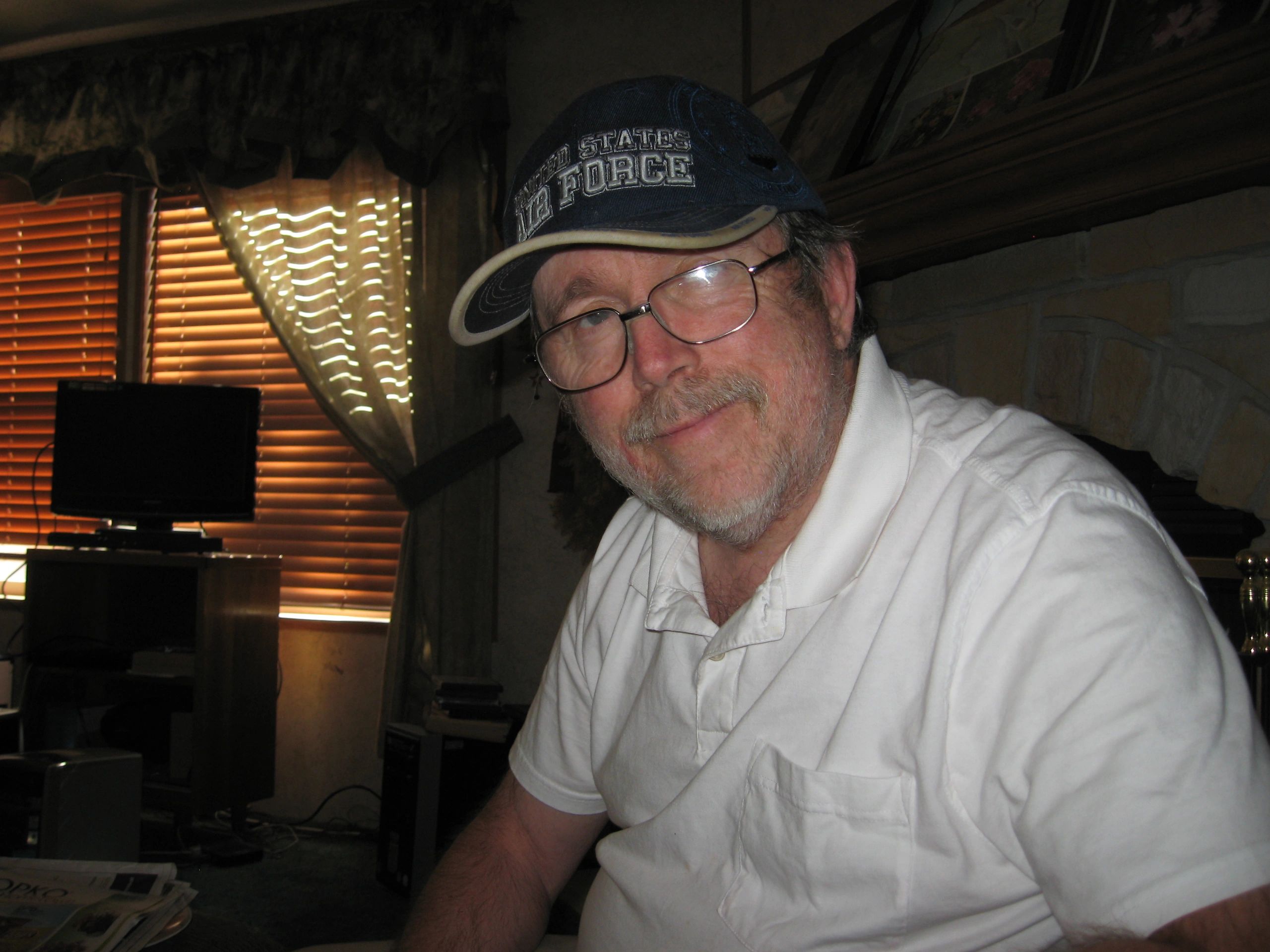 Man in glasses and a USAF cap smiles indoors.
