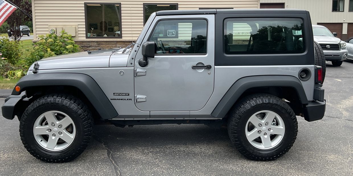 Side view of a silver Jeep Wrangler Sport in a parking lot.