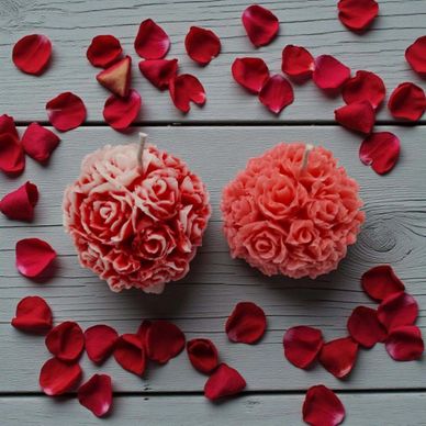 Two rose-shaped candles surrounded by red rose petals on a wooden surface.