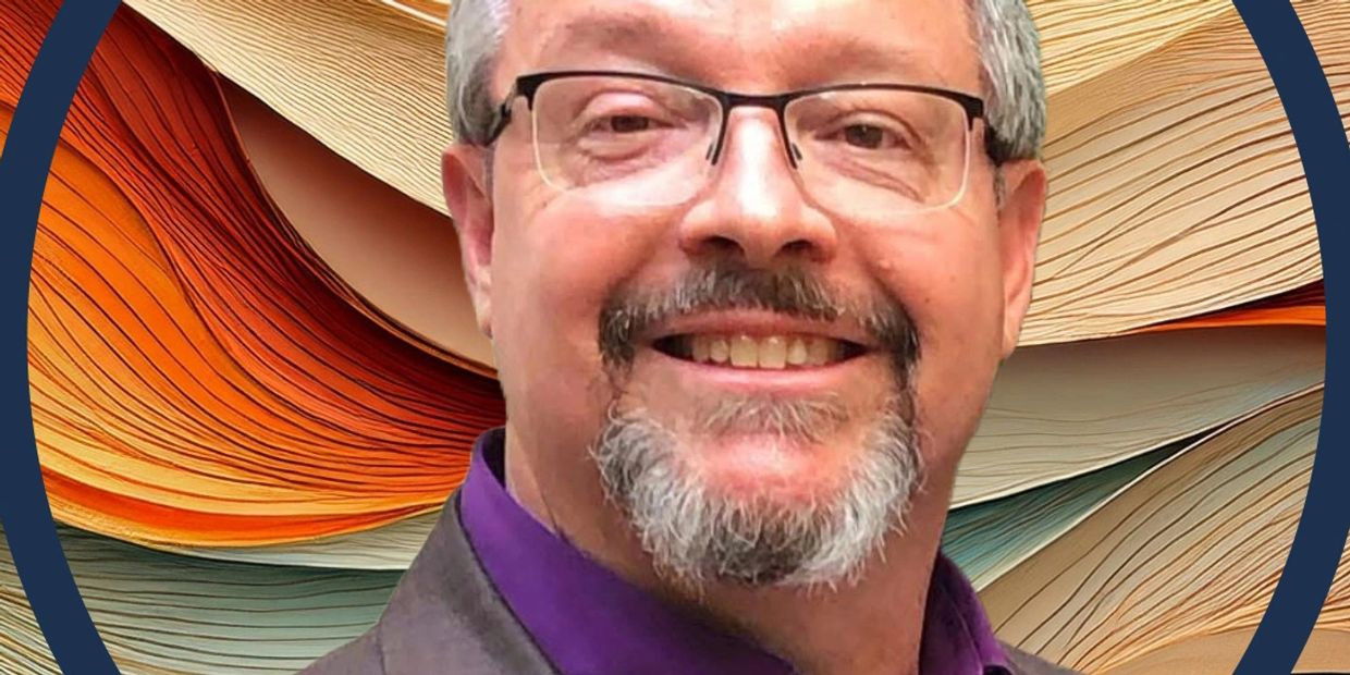 Smiling man with glasses and a gray beard in a purple shirt and brown blazer.