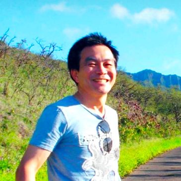 Man smiling outdoors on a sunny day with mountains in the background.