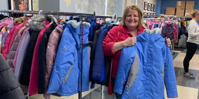 A smiling woman holds a blue coat in a clothing store with racks of jackets.