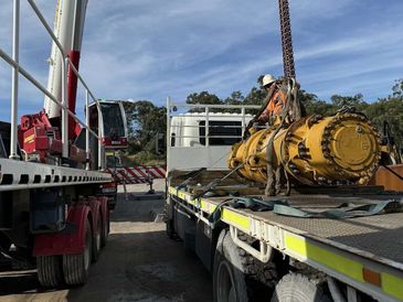 Worker securing large yellow industrial equipment on flatbed truck with crane nearby.