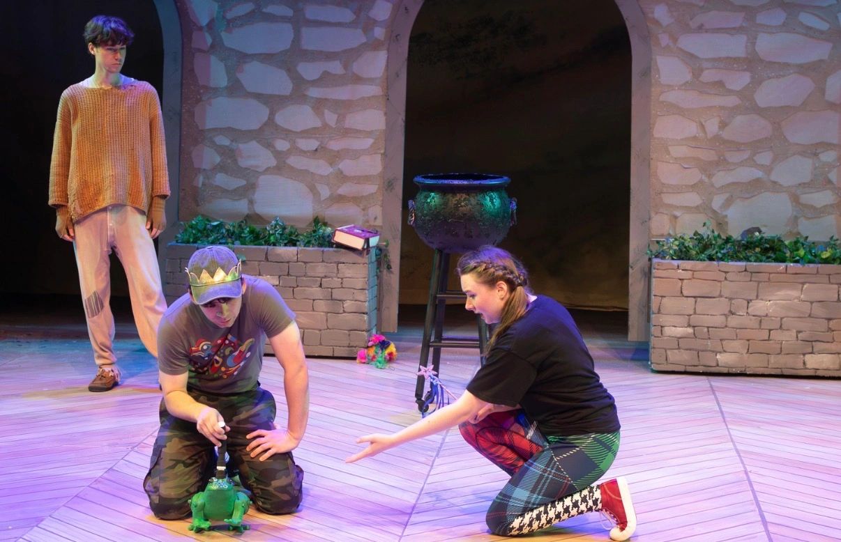 Three people performing on stage with a frog prop.