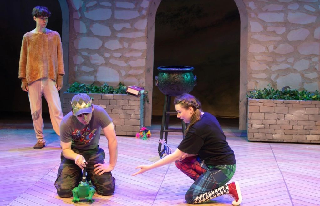 Three people performing on stage with a frog prop.