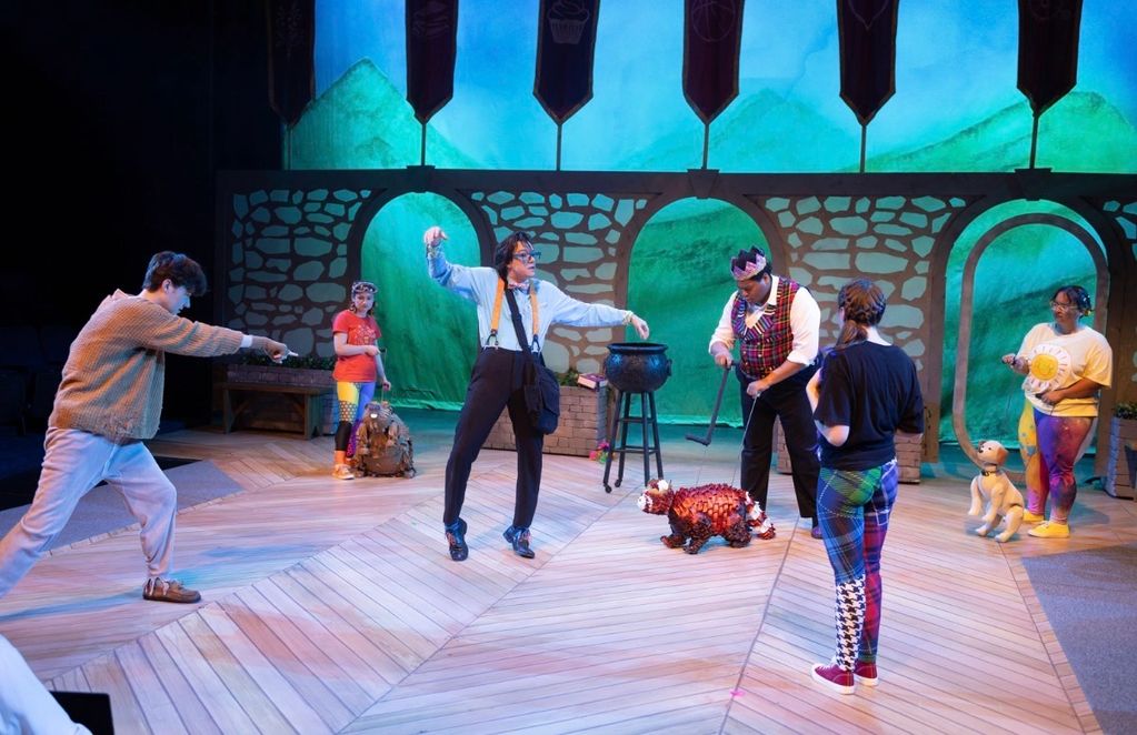 Actors performing on a colorful stage with a dog puppet and a cauldron prop.