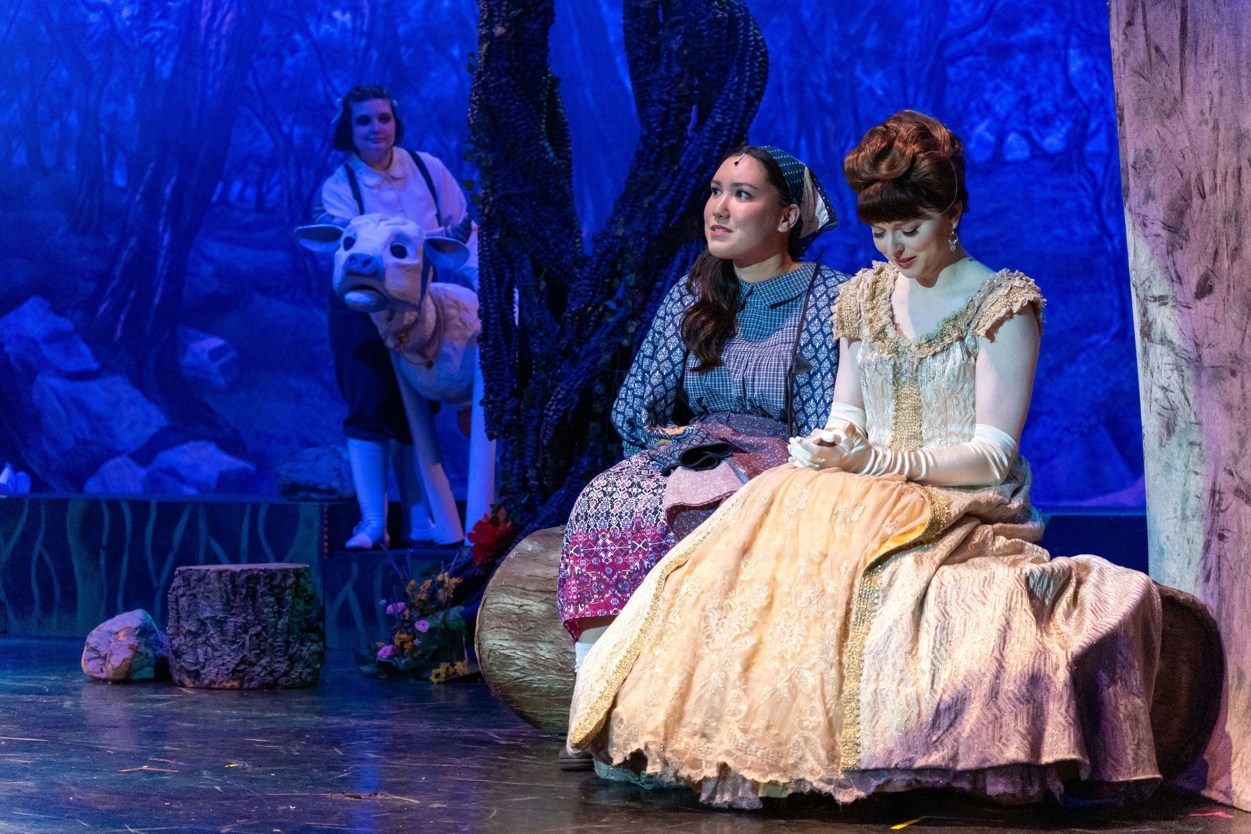 Actors in costume on a theatrical stage with a cow puppet in the background.