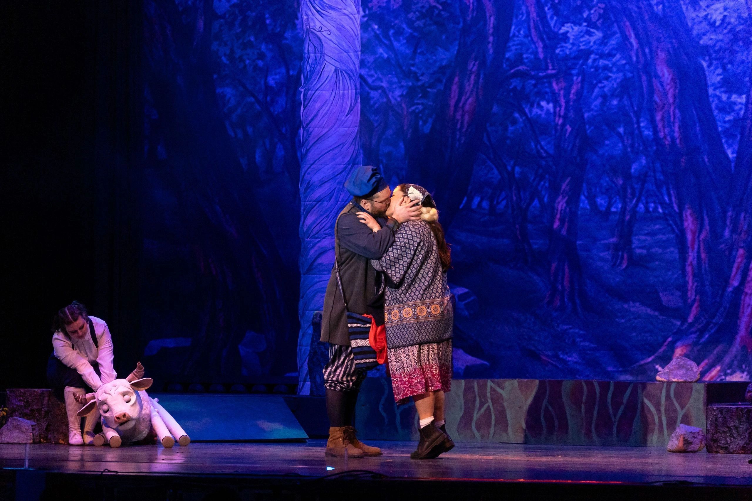 Two actors kissing on stage with a cow puppet behind them.