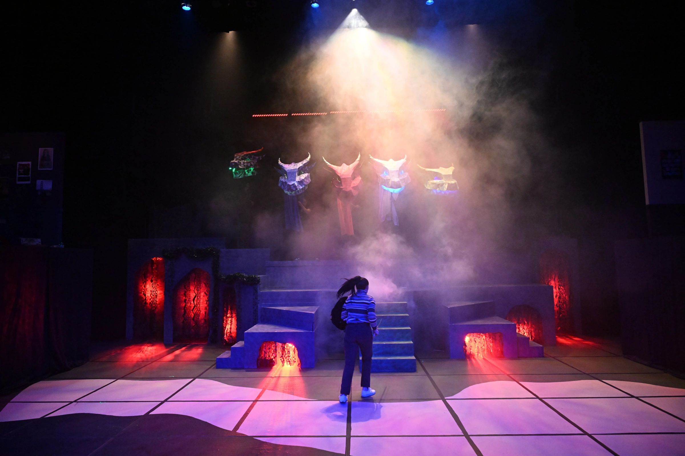 Person under spotlight with costume figures above on a theatrical stage.