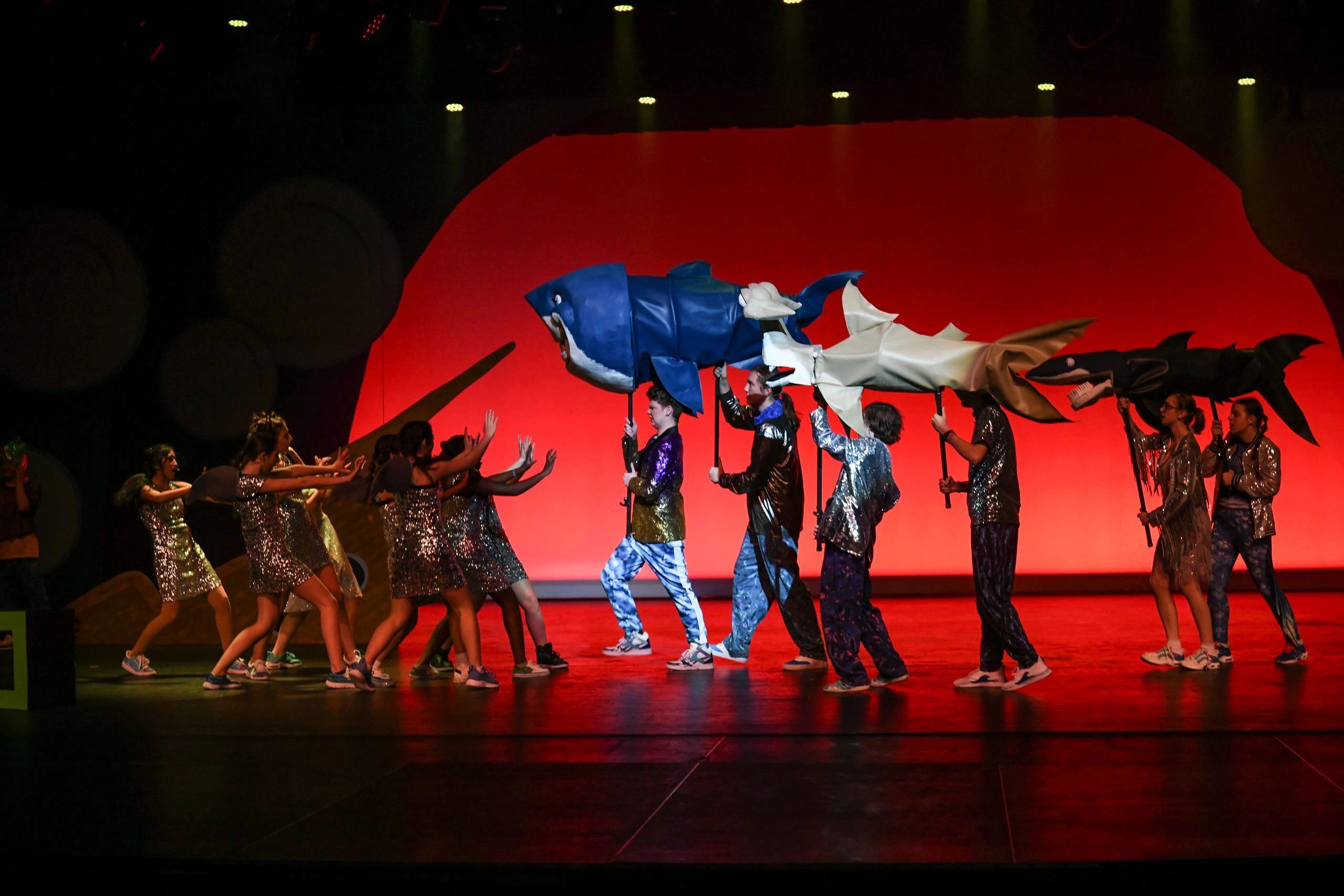 Performers holding large shark puppets on stage with red background.