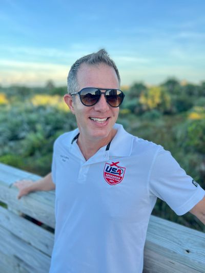 Man wearing sunglasses and a white USA polo shirt, smiling outdoors.
