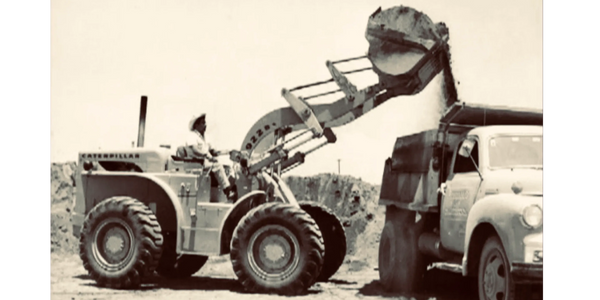 Foto de los 50s de un cargador frontal marca caterpillar vertiendo material en camión de volteo