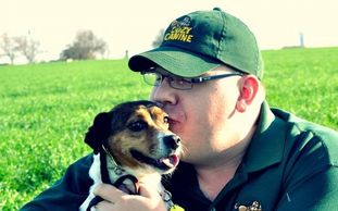 Man wearing a cap kisses a dog outdoors.