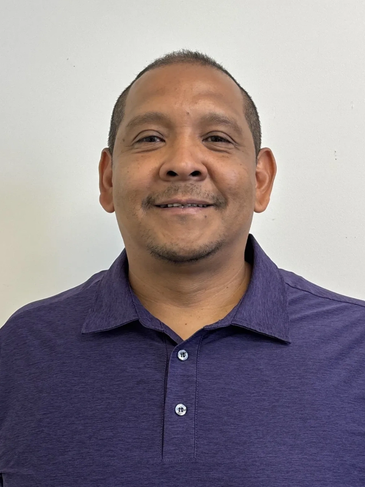 Man smiling in a purple polo shirt against a plain wall.