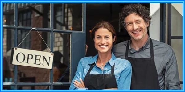 Smiling couple in aprons stands proudly by their open shop door.