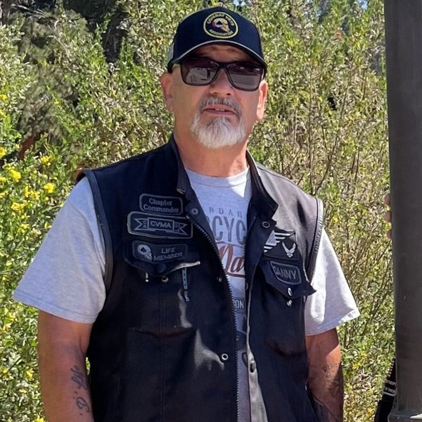 Man wearing a black vest and cap with patches, standing outdoors.