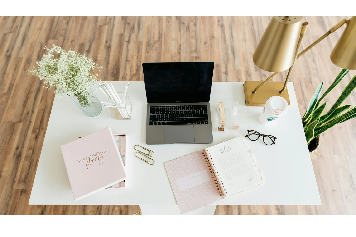 Neat workspace with laptop, planner, flowers, and golden lamp on white desk.
