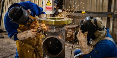 Two workers welding a large metal pipe in an industrial setting with sparks flying.